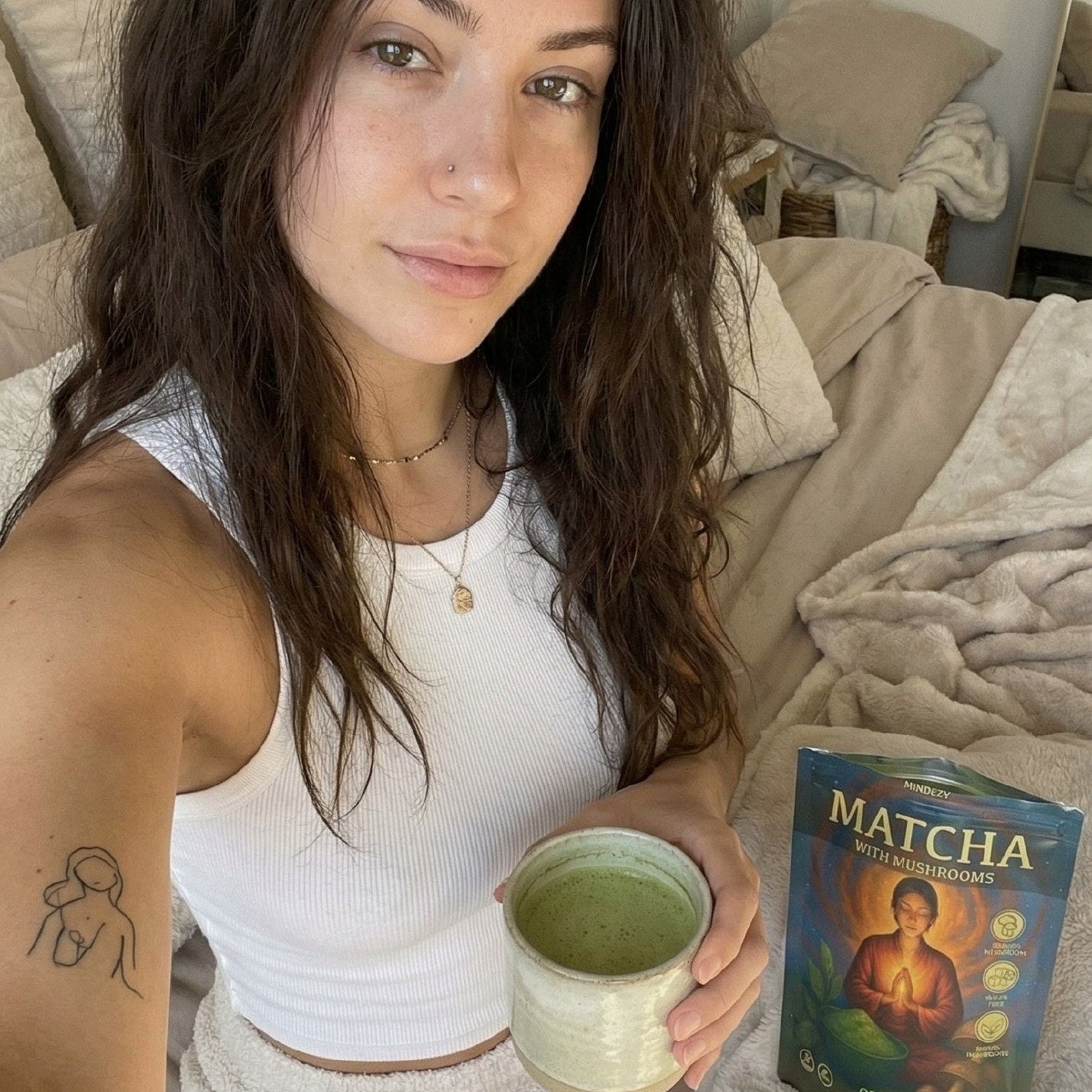 Woman holding a cup of matcha tea with a package of matcha on a bed.